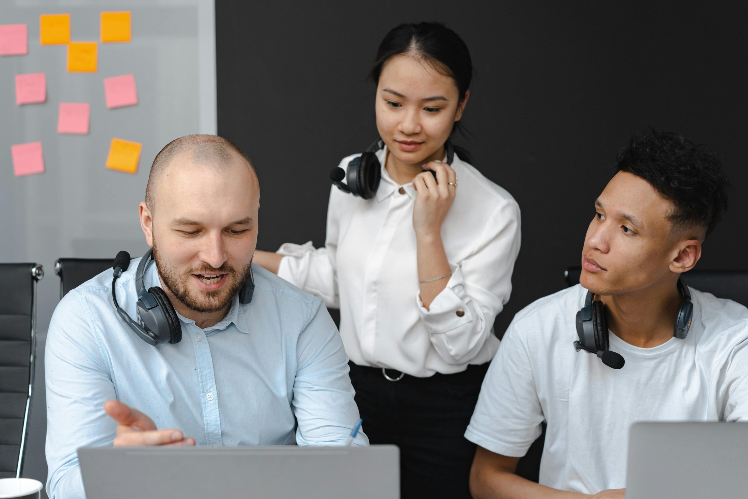 Woman with headphones standing overlooking two colleagues, smiling and demonstrating coaching techniques